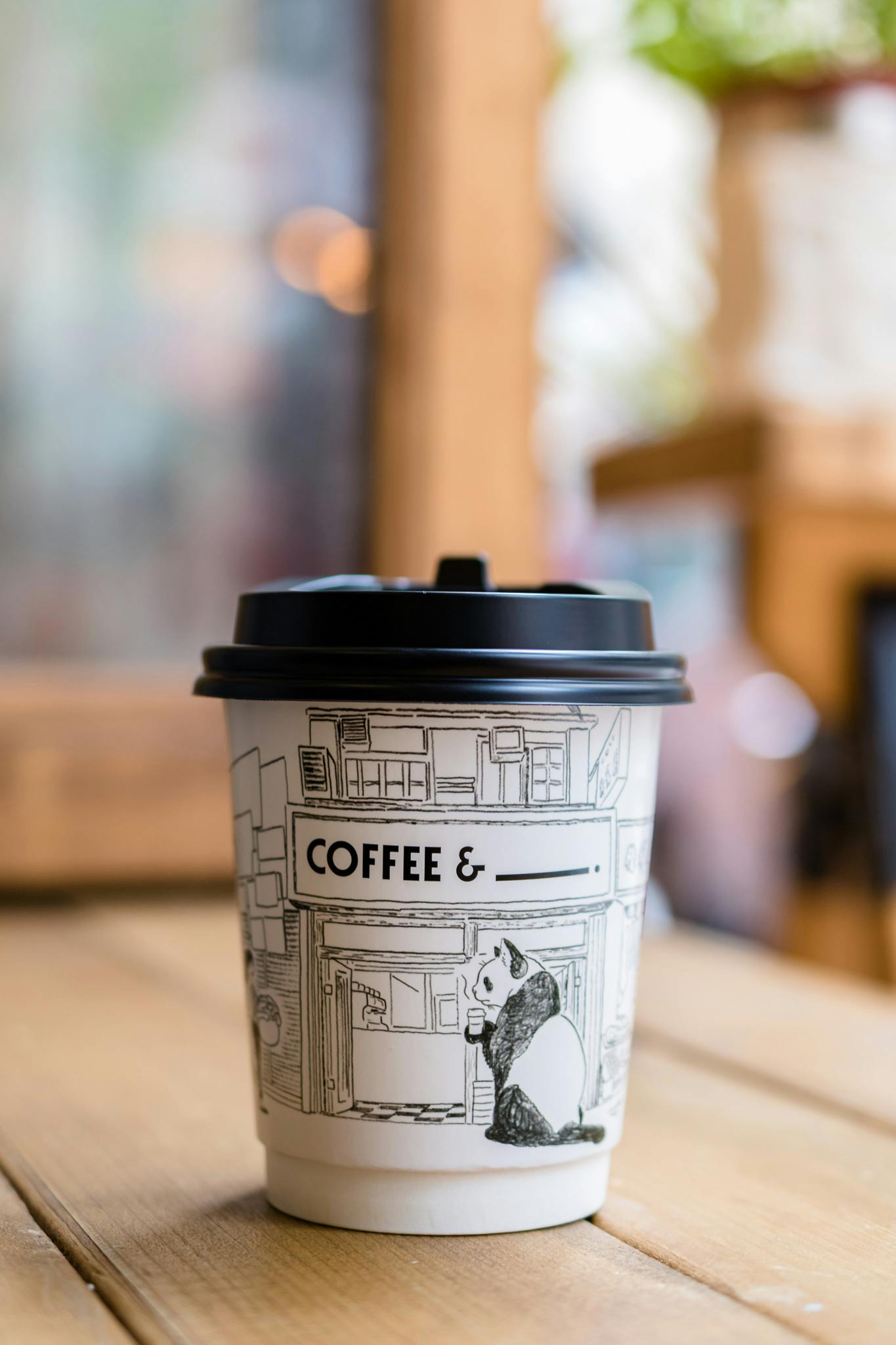 A close-up of a to-go coffee cup with panda art in a cozy cafe setting.